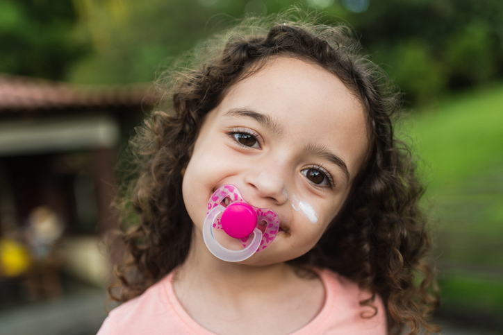 Curly-haired toddler with a pink pacifier in her mouth